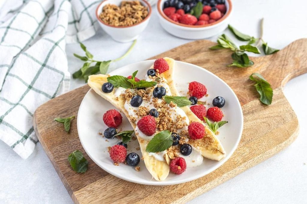 A plate with sliced bananas topped with yogurt, granola, raspberries, blueberries, and mint leaves on a wooden board. Two bowls of granola and berries in the background.