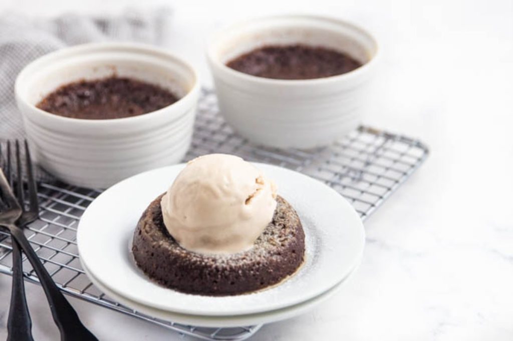 Chocolate lava cake with a scoop of vanilla ice cream on top, served on a white plate. Two additional bowls with chocolate dessert in the background on a cooling rack.