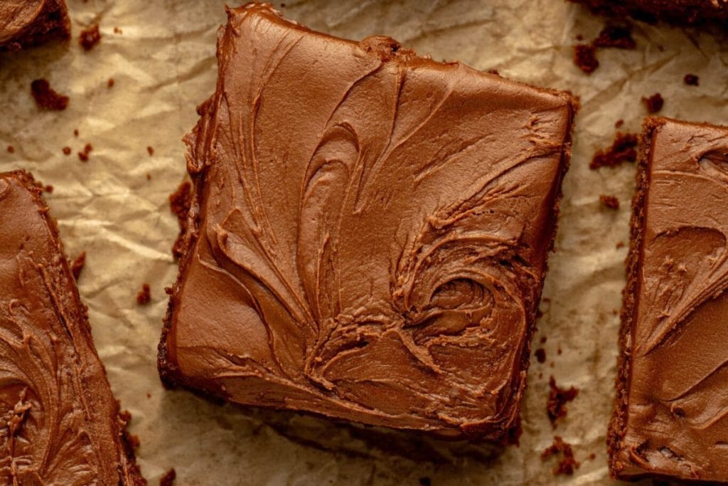 A square piece of chocolate cake with smooth, swirled chocolate frosting sits on crinkled parchment paper.