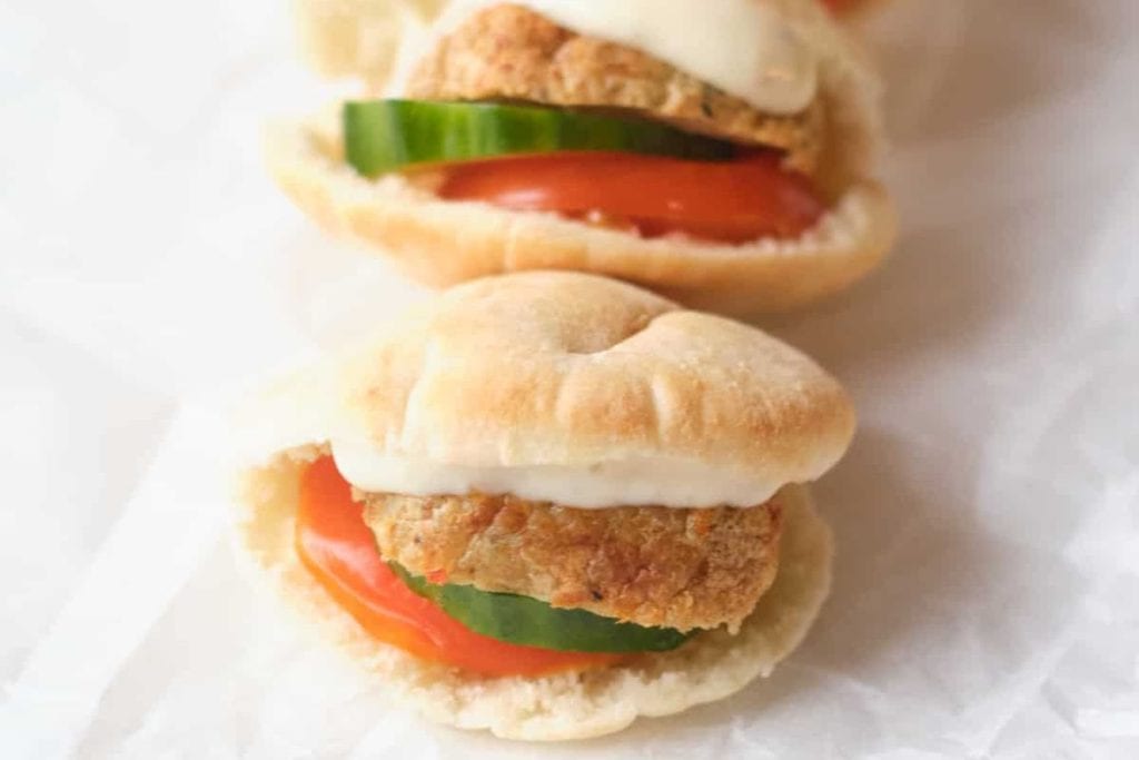 Mini sandwich with fried patty, cucumber slice, tomato slice, and sauce in a small bun on parchment paper.