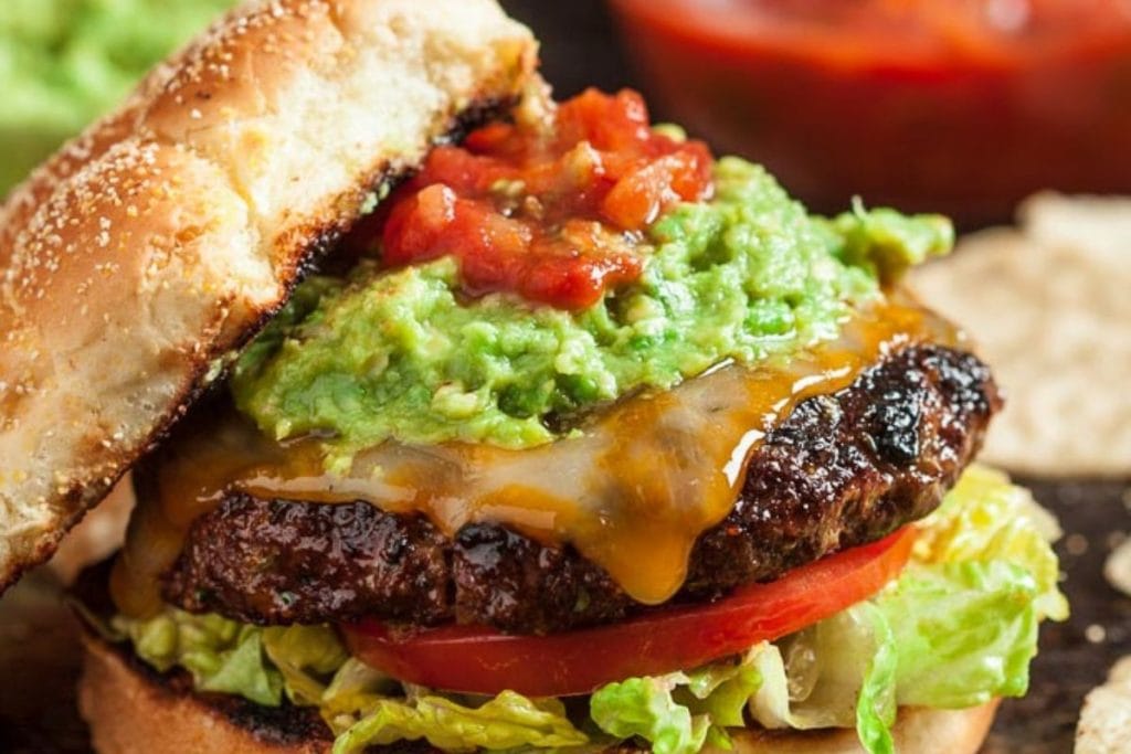 A hamburger with melted cheese, guacamole, and salsa on a sesame seed bun, with lettuce and tomato slices.