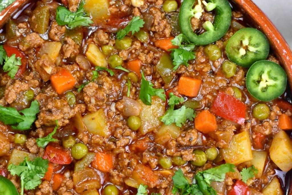 Close-up of a colorful stew with ground meat, diced vegetables, peas, and spices, topped with cilantro and sliced jalapeños.