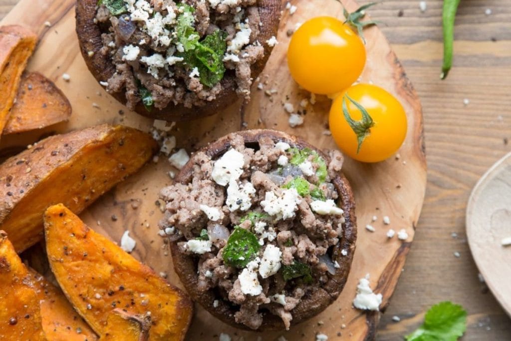 Two stuffed mushrooms with ground meat and herbs, accompanied by sweet potato wedges and two yellow cherry tomatoes, on a wooden surface.