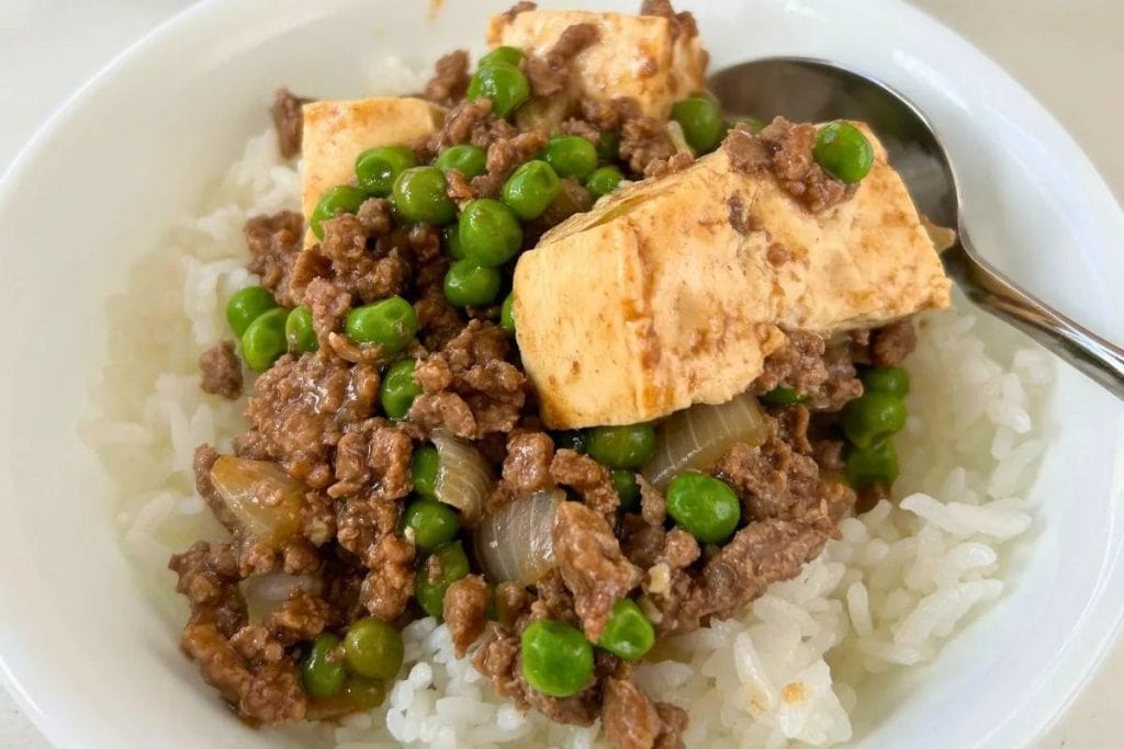 A bowl of white rice topped with ground meat, tofu cubes, green peas, and chopped onions, with a spoon on the side.