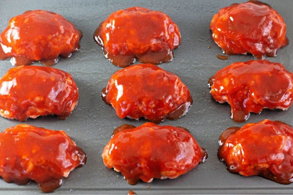 Nine mini meatloaves covered in red glaze on a dark baking tray.