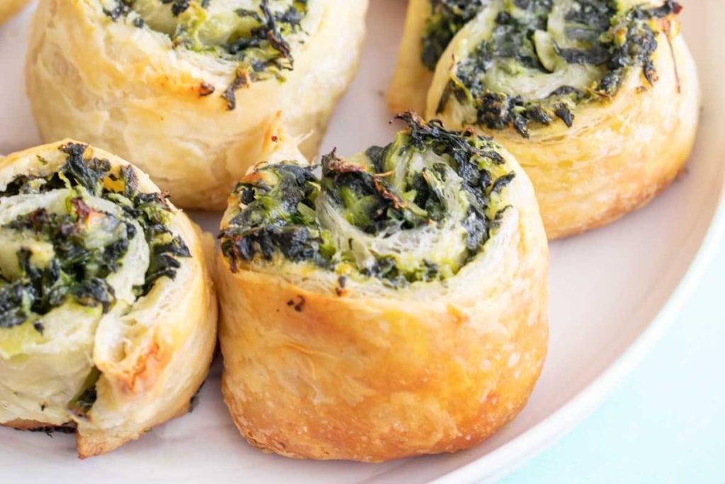Close-up of golden-brown puff pastry pinwheels filled with spinach and cheese, arranged on a white plate.