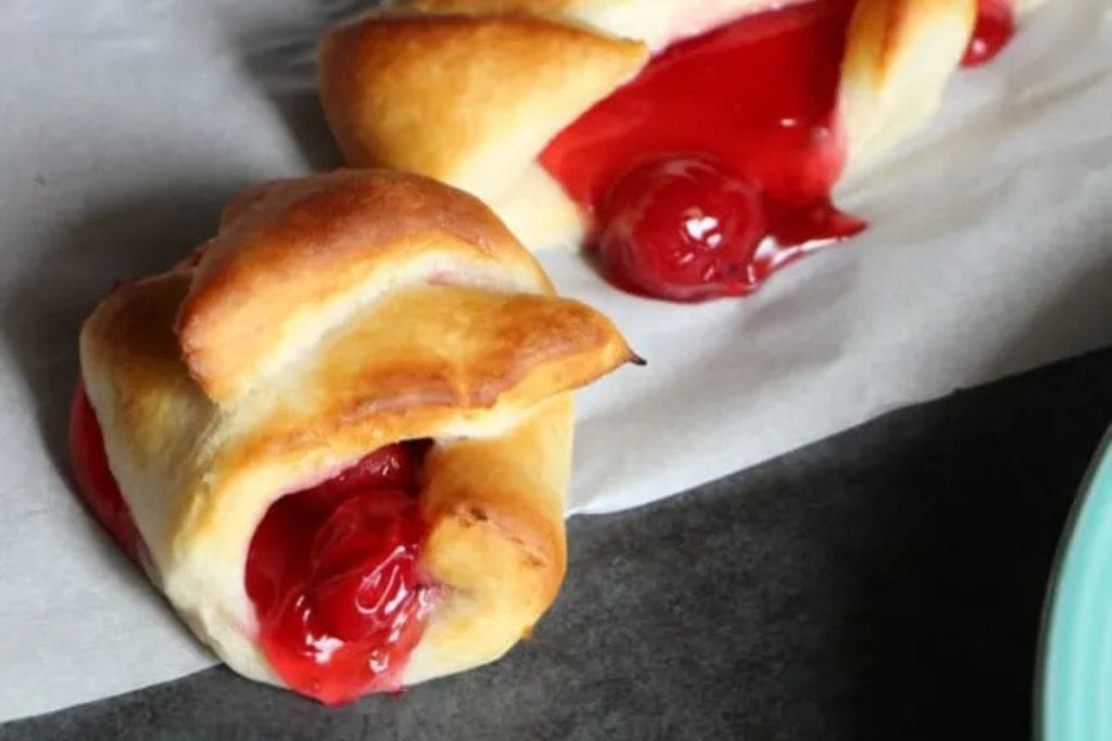 Two baked pastries with golden crusts, filled with bright red cherry filling, resting on parchment paper.