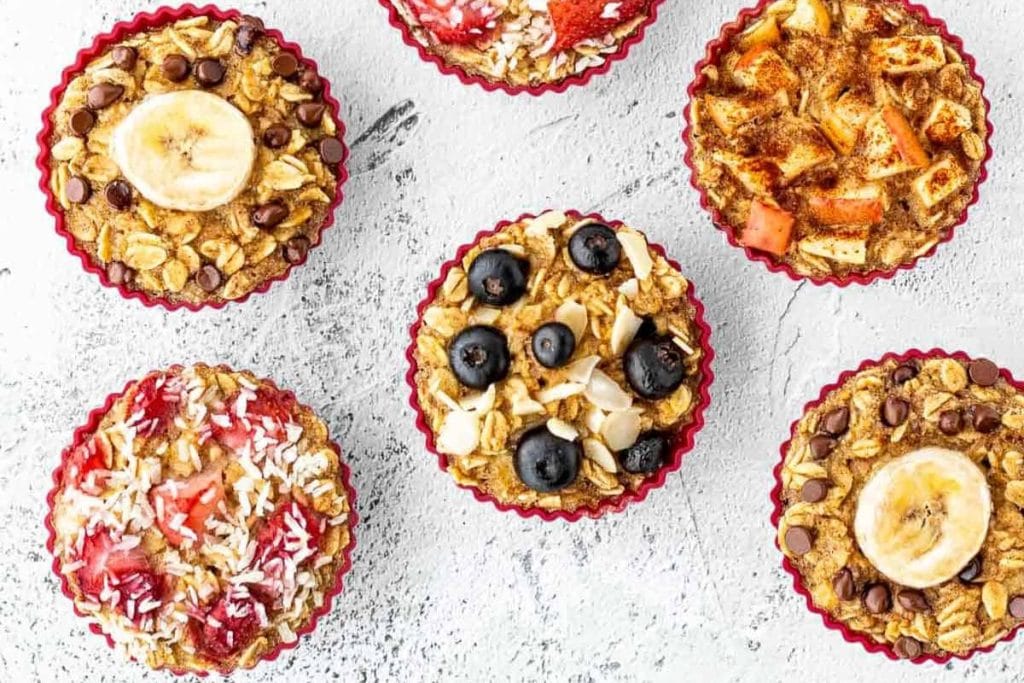 Six baked oatmeal cups in red muffin liners, topped with various ingredients like banana slices, blueberries, strawberries, apple pieces, and chocolate chips, arranged on a textured surface.