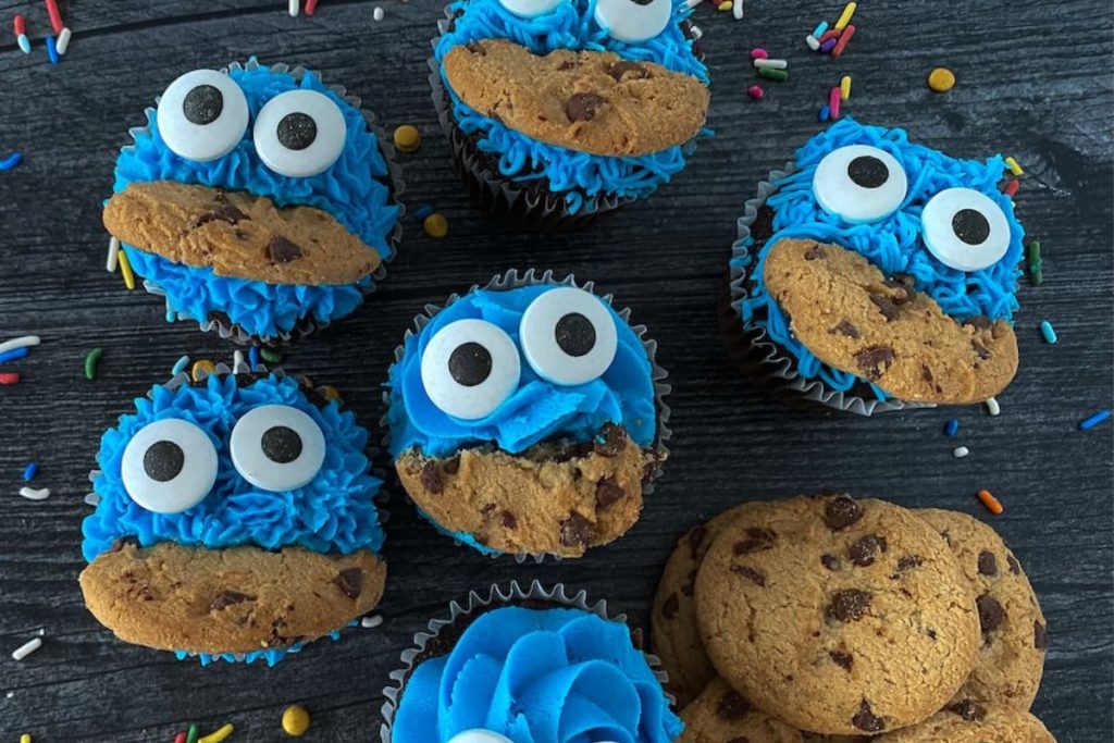 Six cupcakes with blue frosting, large googly eyes, and chocolate chip cookie pieces resembling Cookie Monster, placed on a dark surface with scattered sprinkles.
