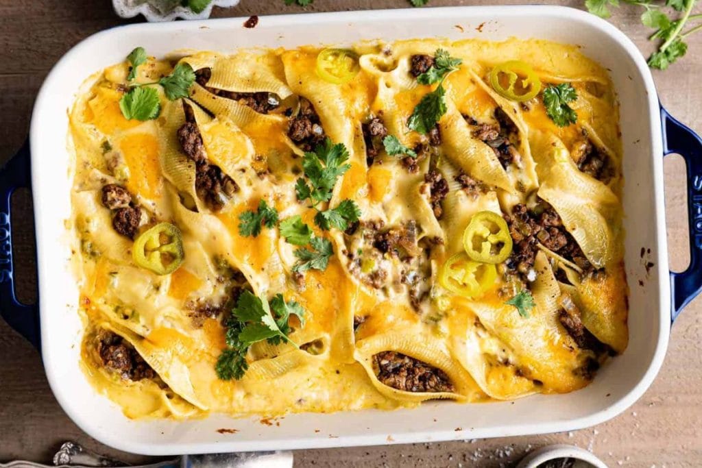 Baked pasta shells filled with meat and cheese, topped with melted cheese, sliced jalapeรฑos, and cilantro in a white rectangular dish.