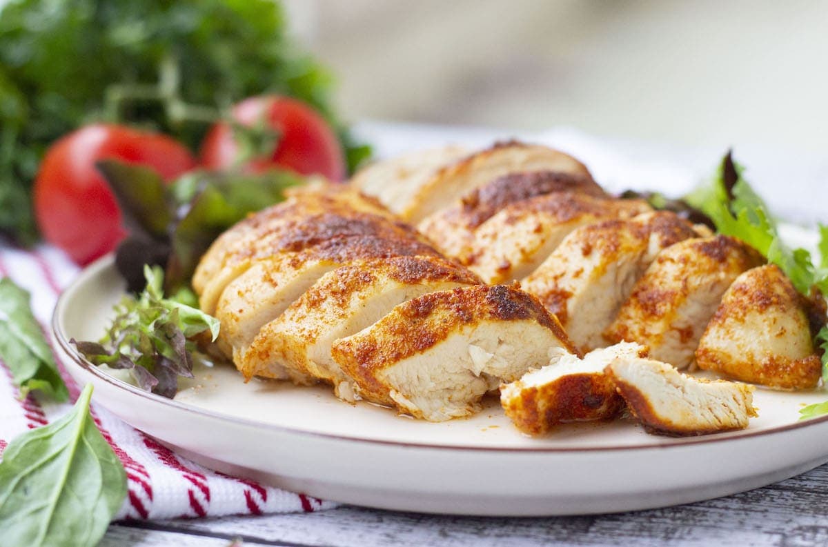 Sliced, seasoned grilled chicken on a plate with mixed greens and tomatoes in the background.