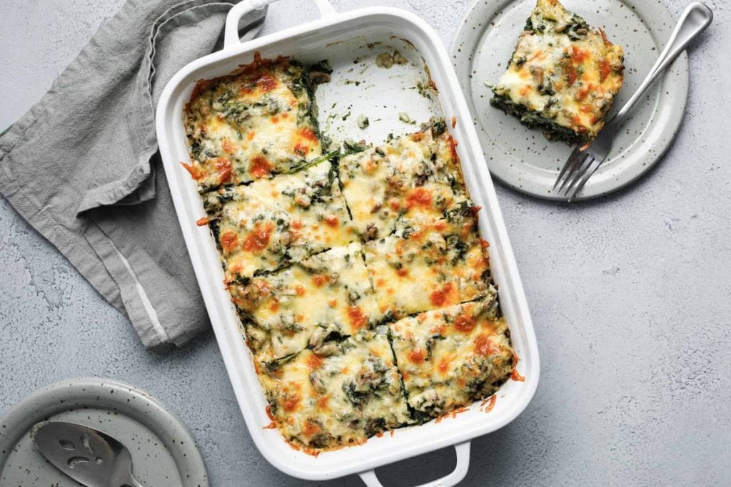 Baked spinach and cheese casserole in a white dish, partially sliced. A serving is on a separate plate with a fork. Gray napkin and serving spatula nearby.