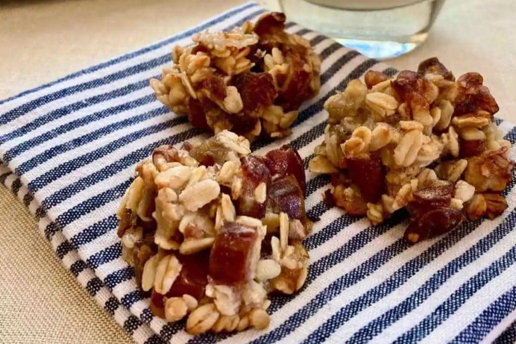 Three oatmeal and date clusters on a striped cloth napkin, with a glass of clear liquid in the background.