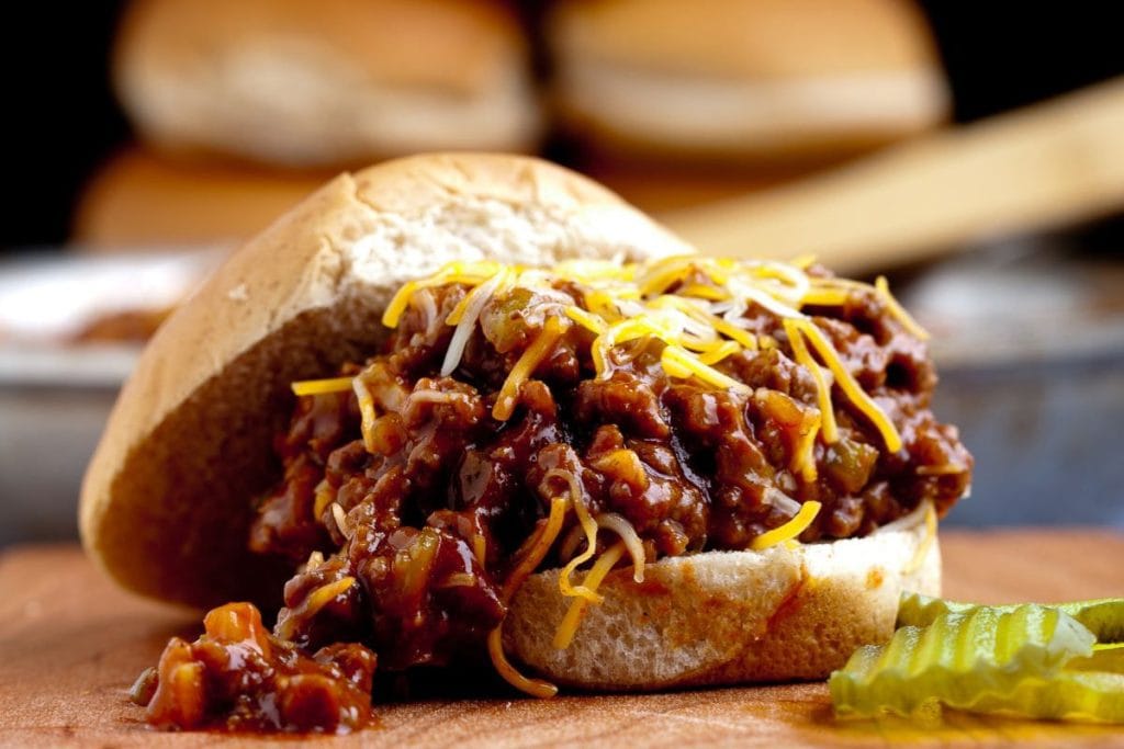 A sloppy joe sandwich with meat sauce and shredded cheese on a bun, accompanied by sliced pickles on a wooden surface.