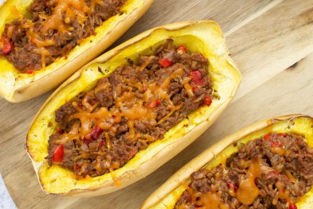 Three spaghetti squash halves filled with ground meat and red peppers, topped with melted cheese, placed on a wooden surface.
