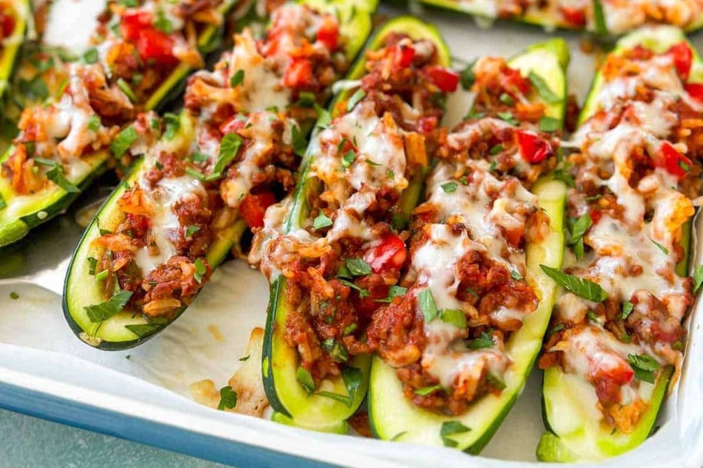 Zucchini boats filled with a mixture of ground meat, diced tomatoes, herbs, and melted cheese, placed on a baking tray.