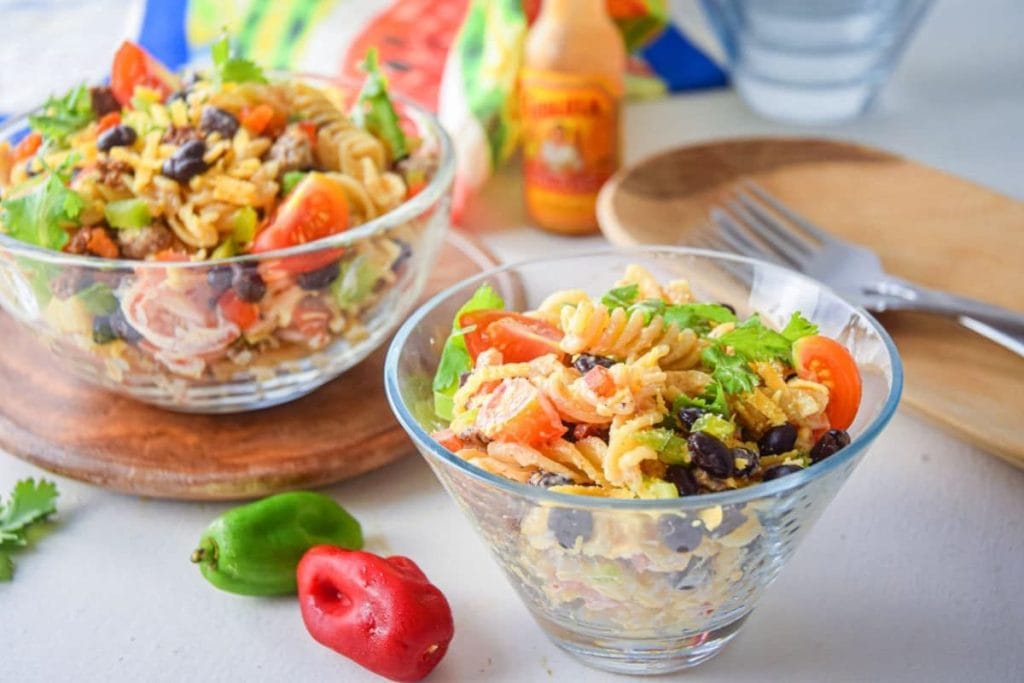 Two bowls of pasta salad with cherry tomatoes, black beans, and lettuce. A bottle of hot sauce and various peppers are nearby on a white surface.