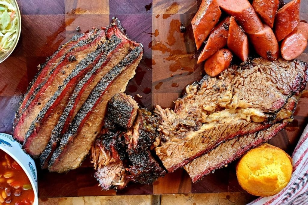 Slices of brisket and sausage on a wooden board with a serving of cornbread on the side.