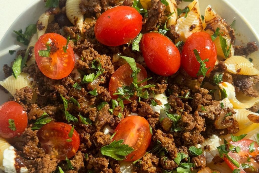 A dish of pasta shells topped with ground meat, halved cherry tomatoes, chopped herbs, and sliced onions.