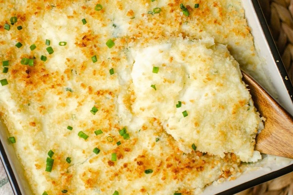 A baking dish with golden-brown mashed potatoes topped with breadcrumbs and chopped chives. A wooden spoon is scooping a portion from the side.