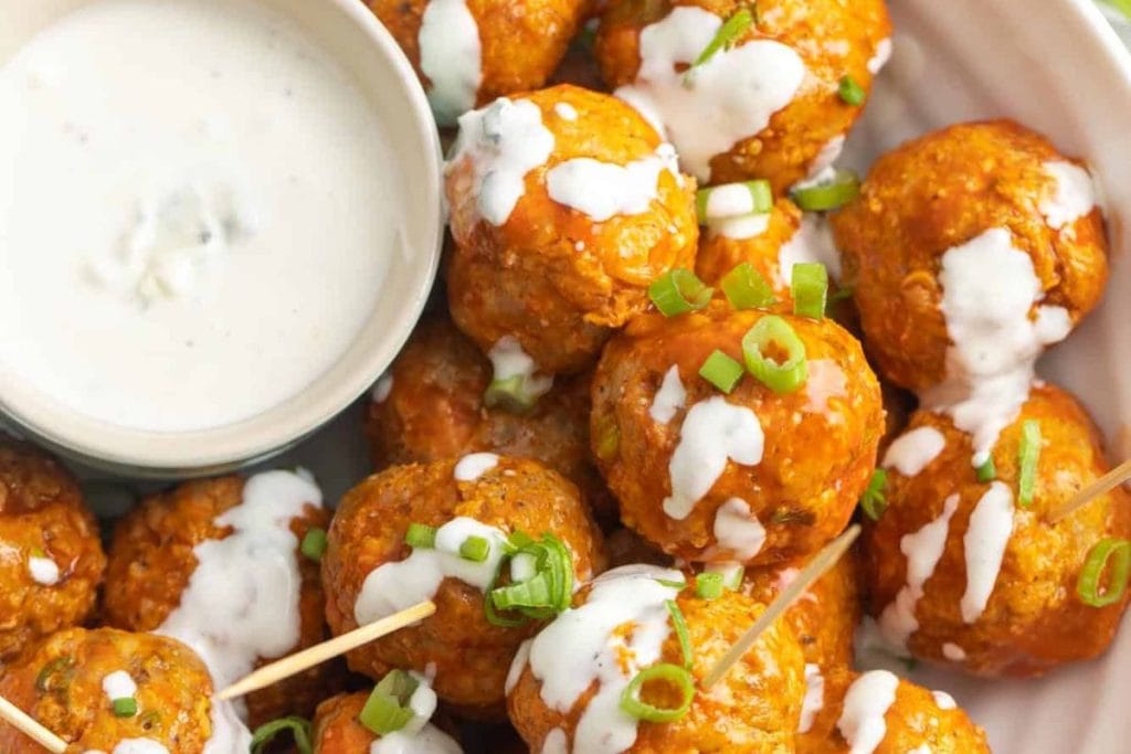 Buffalo chicken meatballs drizzled with white sauce, garnished with chopped green onions, served with a small bowl of dipping sauce.