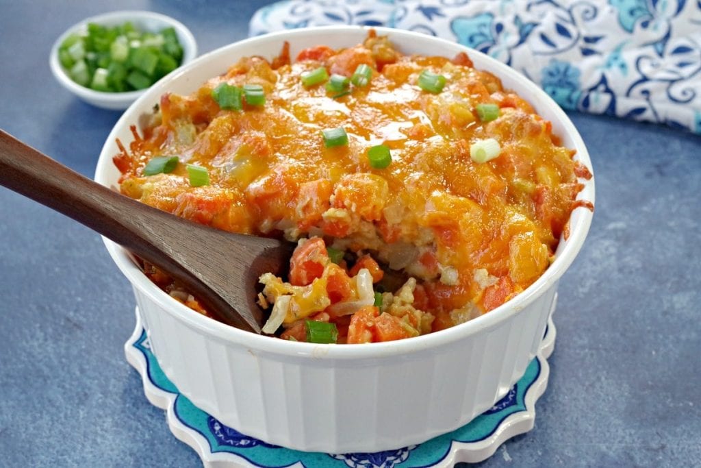 A cheesy casserole topped with green onions in a white dish, with a wooden spoon. A small bowl of chopped green onions is in the background.