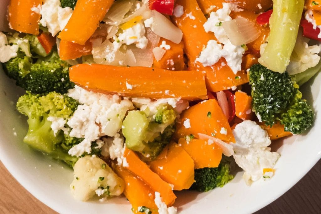 A white bowl filled with a colorful mix of chopped carrots, broccoli, cauliflower, red peppers, onions, and feta cheese.
