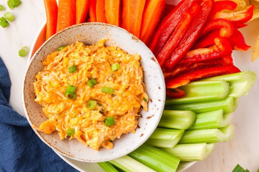 A bowl of buffalo chicken dip garnished with green onions, surrounded by carrot sticks, red bell pepper slices, and celery sticks.