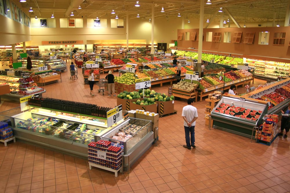 A spacious grocery store interior with various sections displaying fresh produce, packaged goods, and a bakery. Shoppers are browsing different aisles under bright lights and high ceilings.