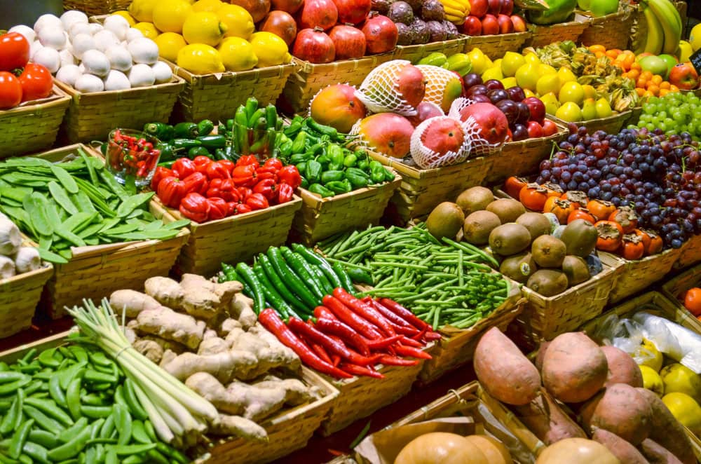 Baskets of various fruits and vegetables, including tomatoes, peppers, beans, ginger, kiwis, grapes, and mangoes, are neatly arranged at a market stall.