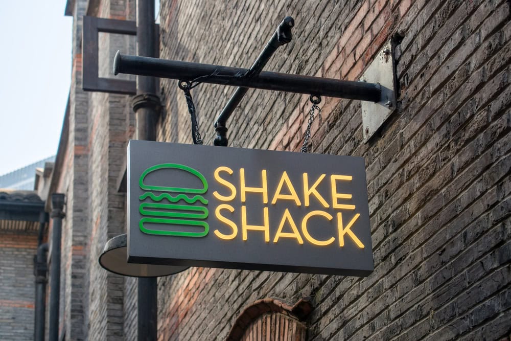 Sign for Shake Shack hangs on a brick wall, featuring a green burger logo and yellow text.