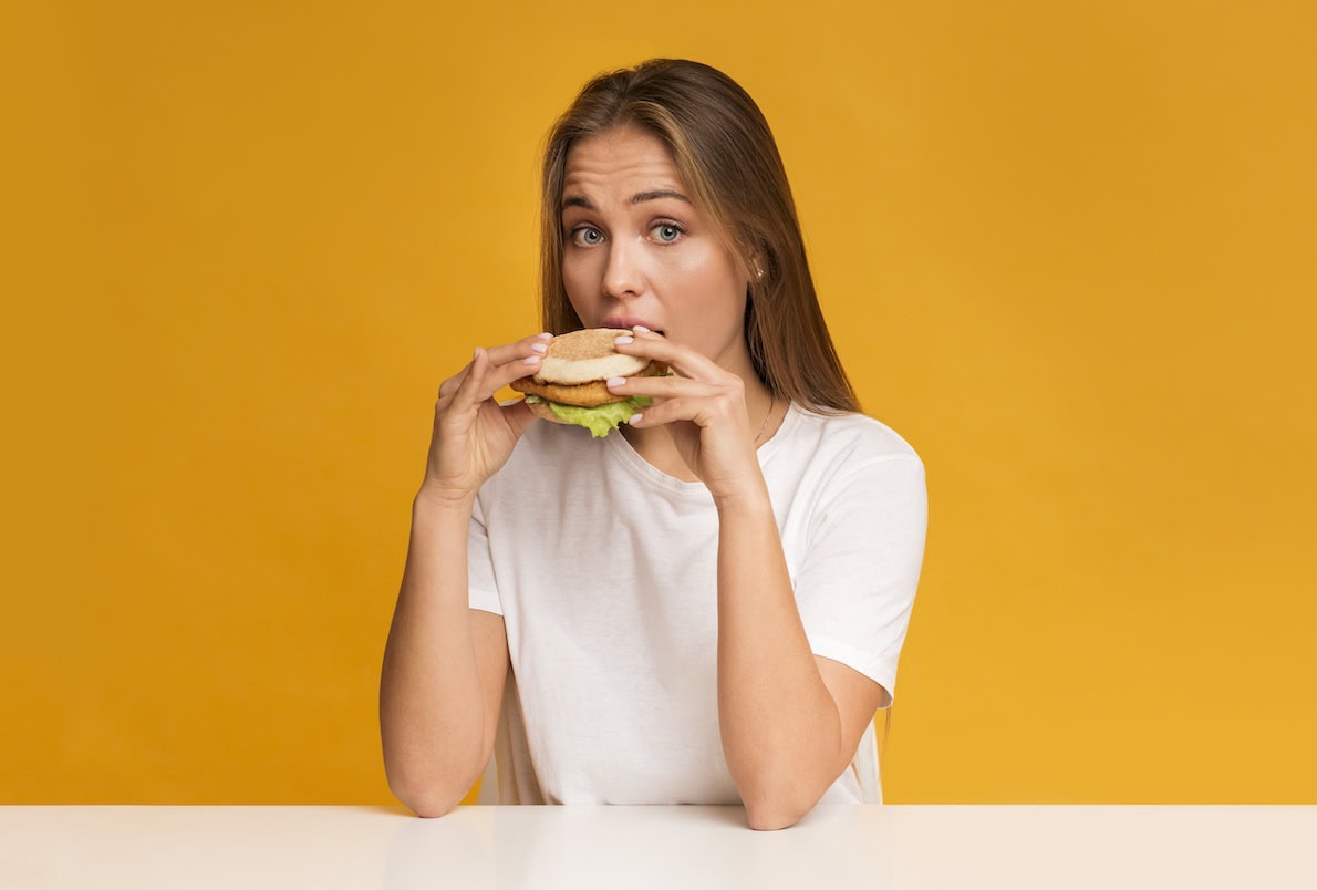 Person in a white shirt eating a burger against a solid yellow background.