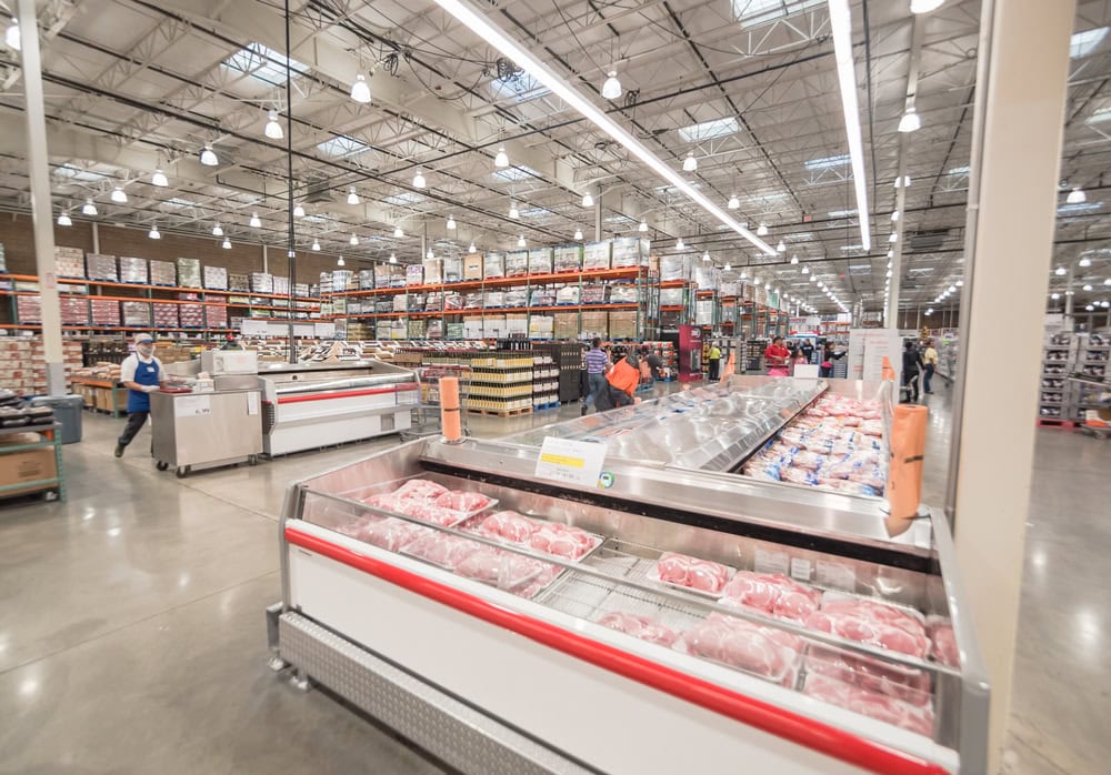 Interior of a large warehouse store, featuring wide aisles, high shelves stocked with goods, and refrigerated sections with meat. Shoppers and employees can be seen throughout.