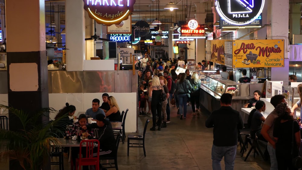 People dining and walking in a busy indoor food market with various food stalls and neon signs.