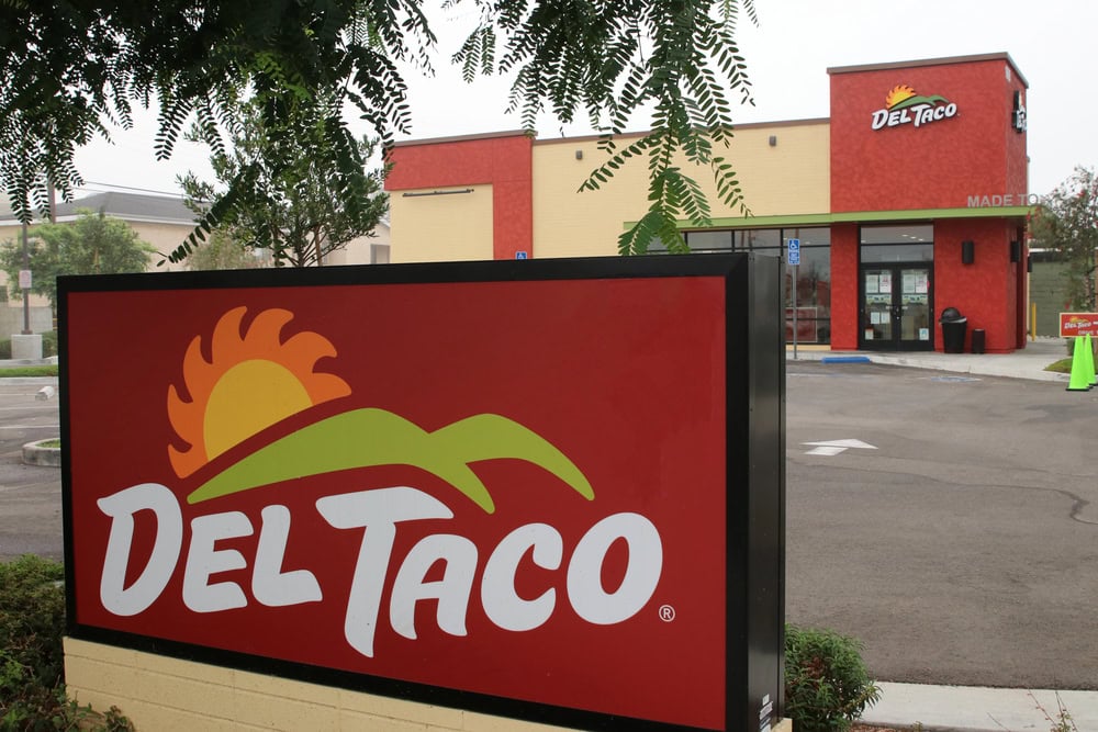 Del Taco restaurant exterior with large sign featuring the logo, surrounded by trees and an empty parking lot.