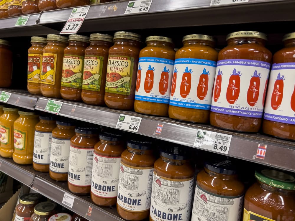 Shelves filled with various brands of pasta sauce jars, including Classico and Carbone, with visible price tags in a grocery store aisle.