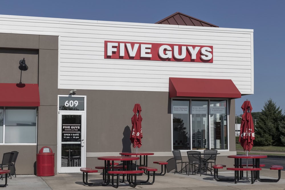 Outdoor view of a Five Guys restaurant with red accents, umbrellas, and seating. A door sign shows open hours from 11 a.m. to 9 p.m. Clear sky in the background.