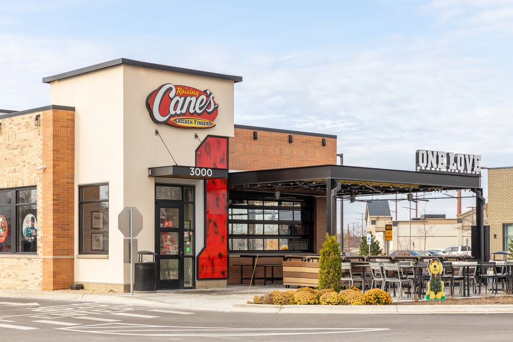Restaurant exterior with beige and brick facade, featuring "Raising Cane's Chicken Fingers" signage. "One Love" sign on adjacent patio with outdoor seating and greenery.