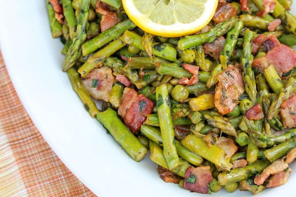 Plate of sautéed asparagus and peas mixed with bacon pieces, garnished with a lemon slice, on a white plate.