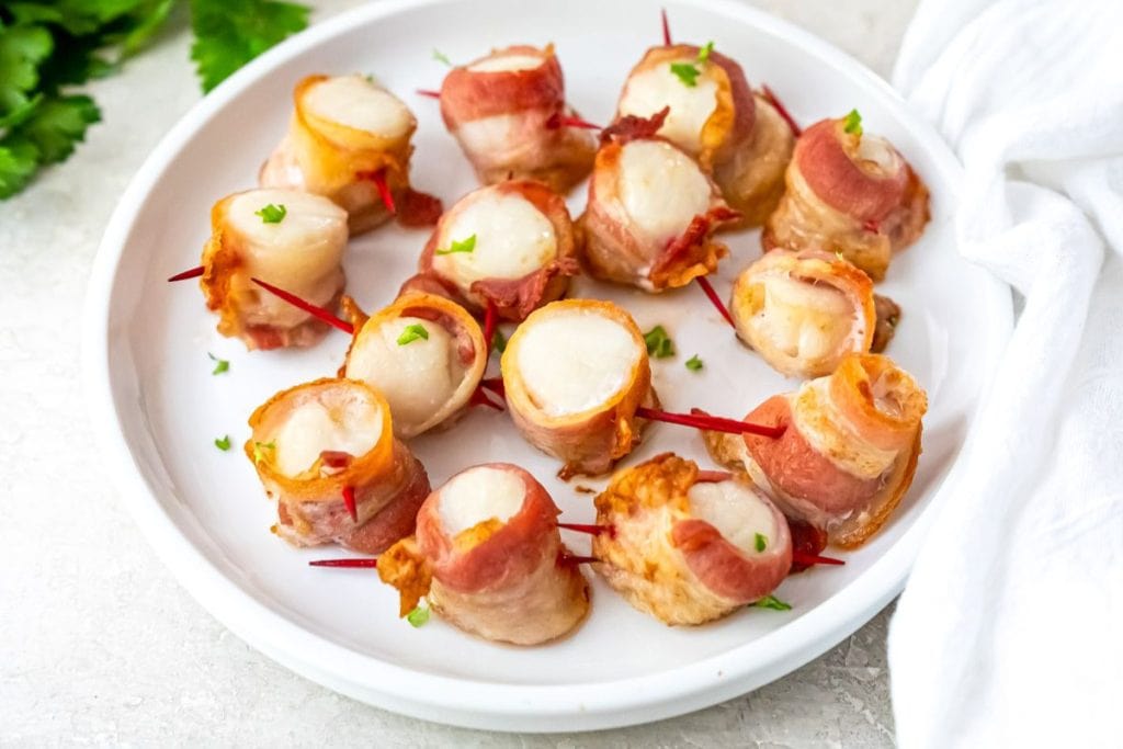 A plate of bacon-wrapped scallops, garnished with chopped herbs and secured with toothpicks, on a light-colored surface.
