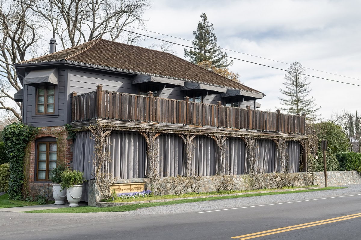 Two-story building with dark siding and wooden balcony. Curtains hang on the lower level, and there's a small sign near a bench. Trees and a road are visible in the background.
