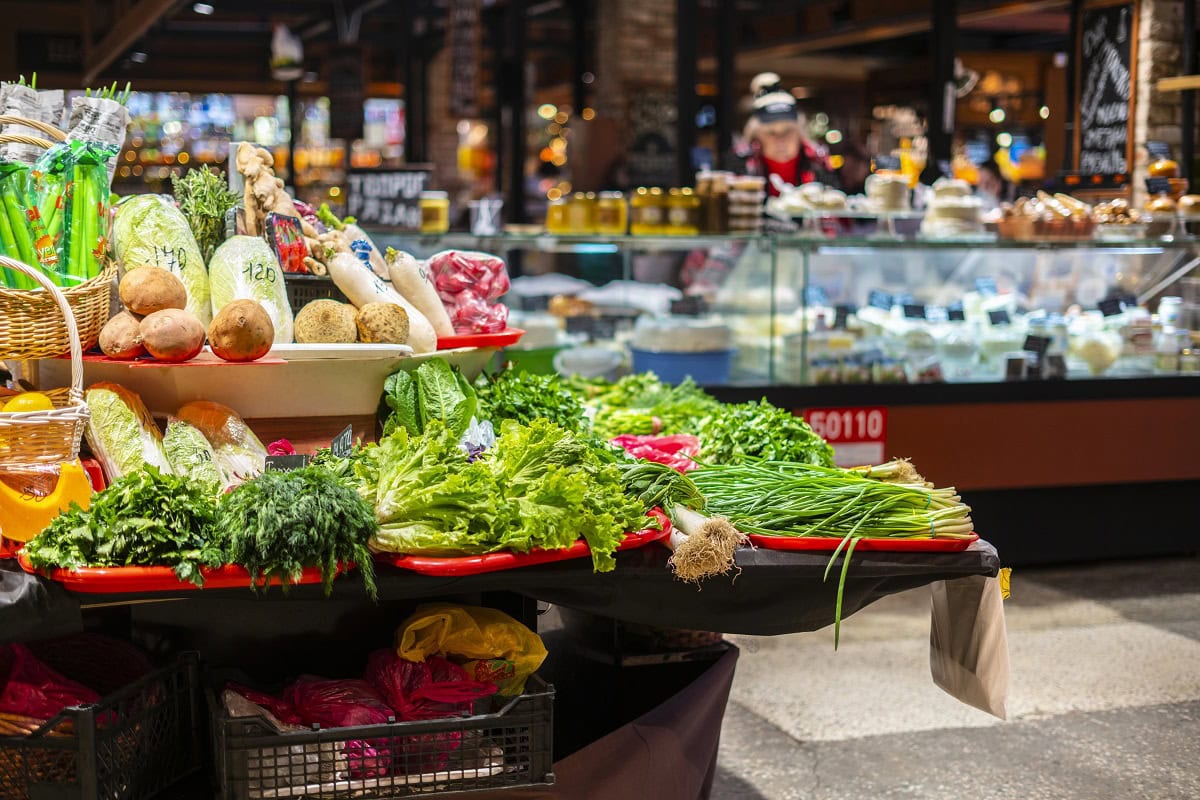 A market stall displays fresh vegetables, including greens and radishes, with a deli counter visible in the background.