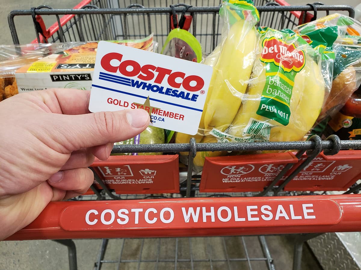 Hand holding a Costco Wholesale Gold Star Member card in front of a shopping cart filled with groceries, including bananas and snacks, at Costco.