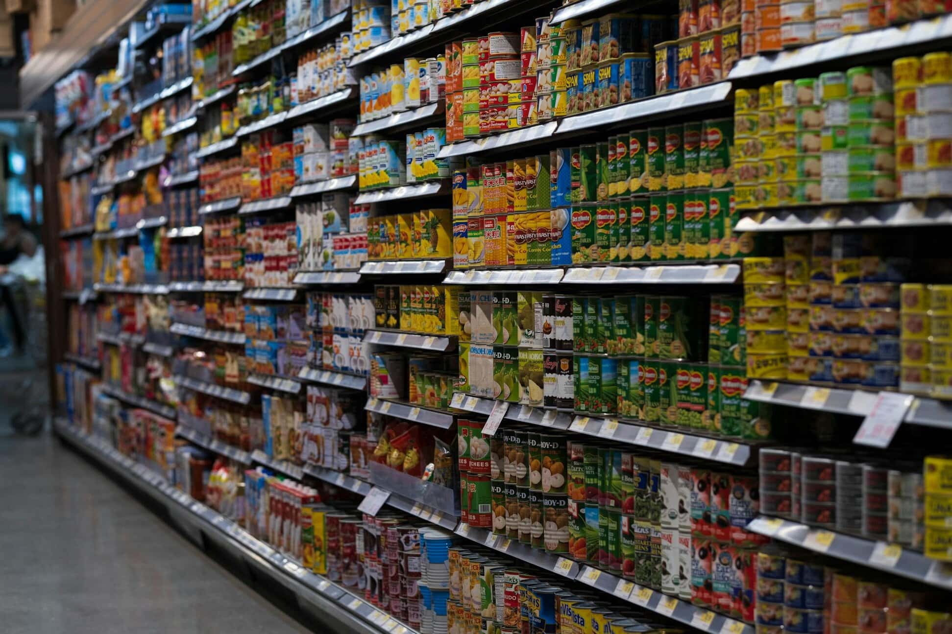 A supermarket aisle filled with neatly arranged canned and packaged goods on shelves, including various brands and colorful labels, with price tags visible beneath each item.