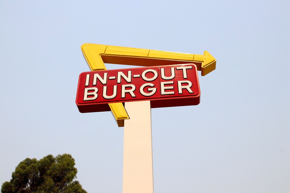 In-N-Out Burger sign with a yellow arrow on a tall pole, set against a clear sky. Trees are partially visible on the left side.