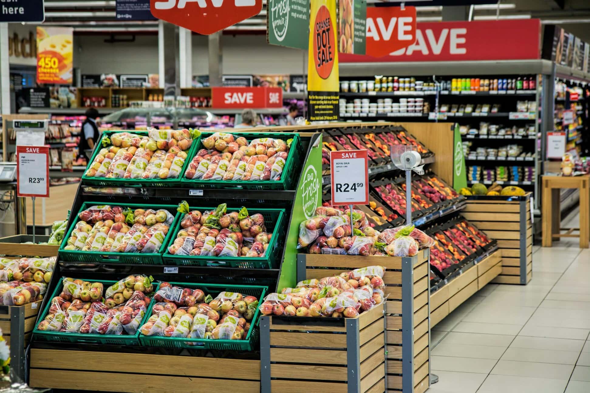 Supermarket aisle with fresh produce displays including apples and oranges. Price tags are visible. Shelves in the background contain various products.