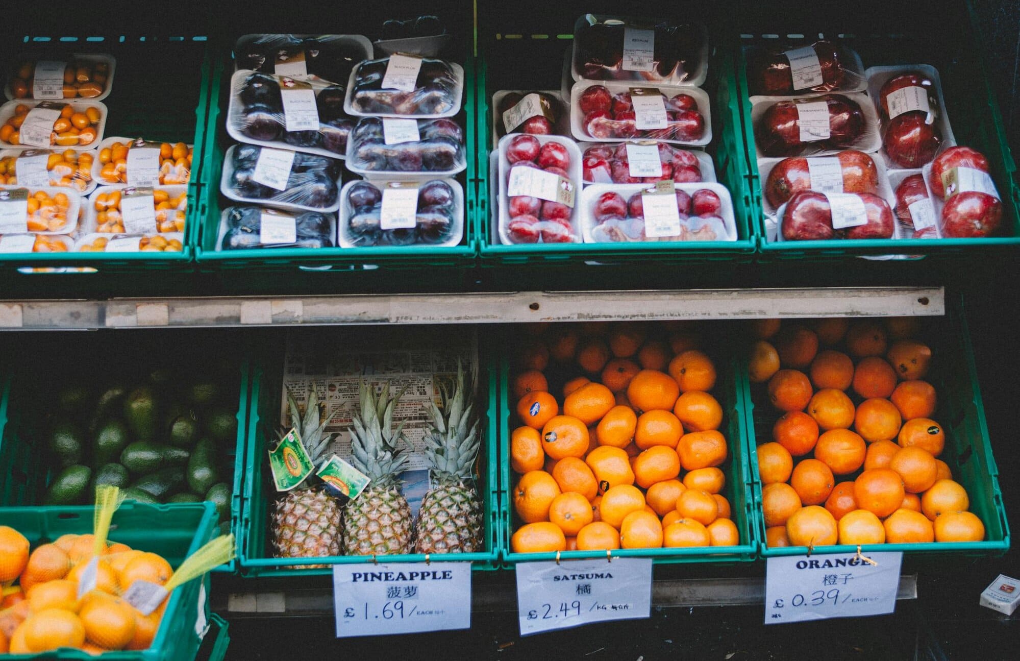 Fruit stand display with packaged plums, apples, and loose avocados, pineapples, satsumas, and oranges. Price labels in British pounds visible for each fruit type.