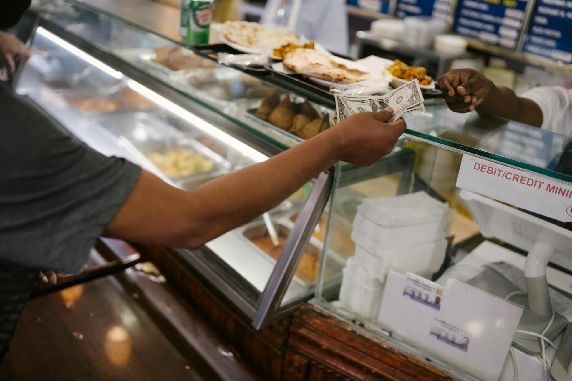 A person hands cash to a cashier across a food counter, with various dishes and a debit/credit sign visible.
