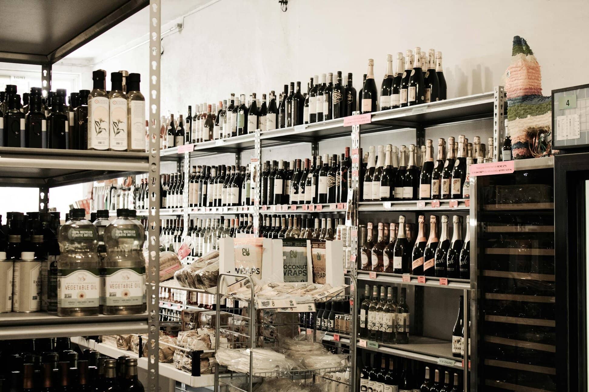 Store interior with shelves displaying various wine bottles, liquors, and other beverages. A small selection of snacks is on the central rack.