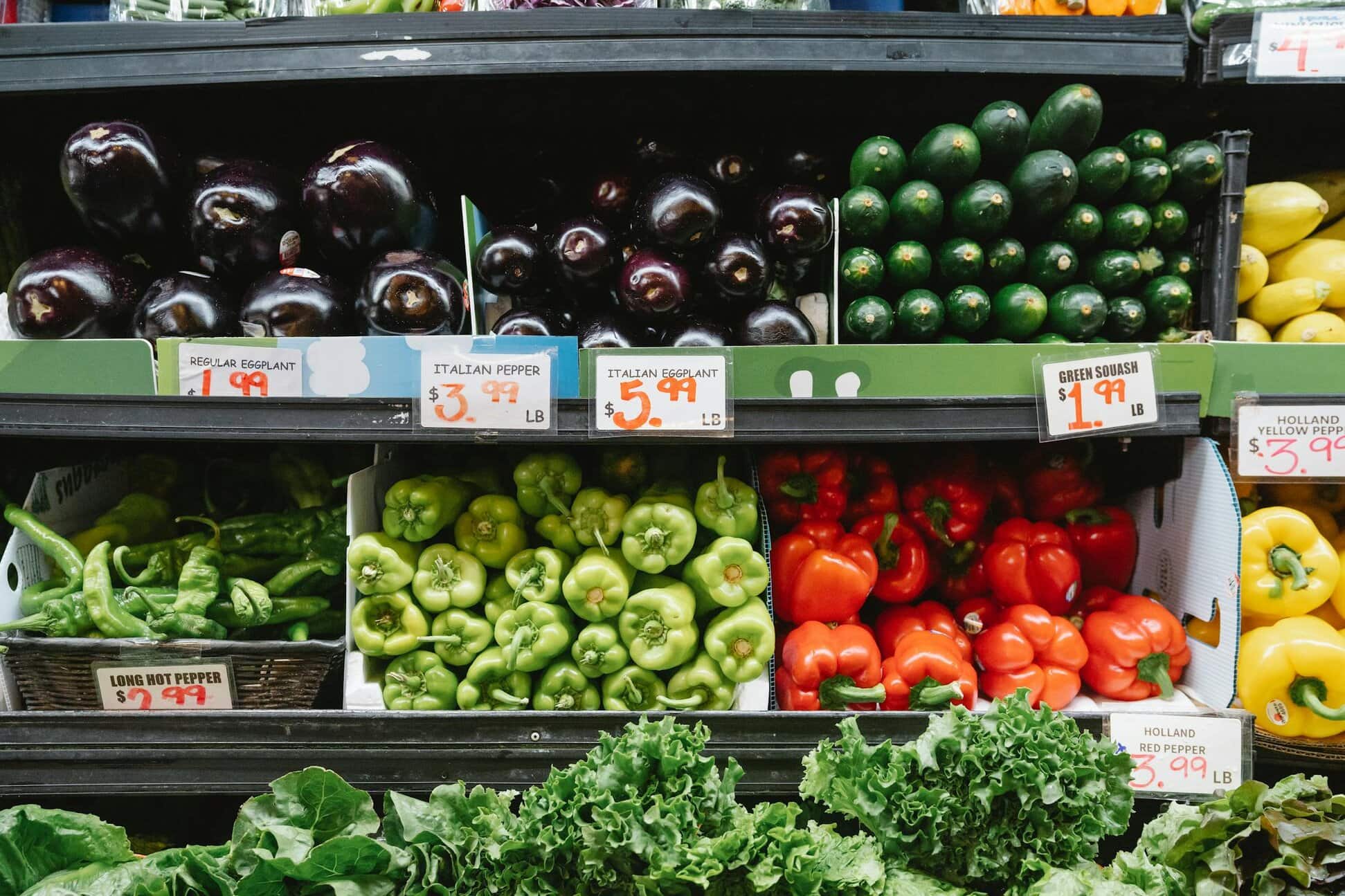 Assortment of vegetables displayed in a grocery store, including eggplants, cucumbers, various peppers, and leafy greens, with visible price tags.